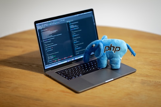 A laptop displaying code on its screen sits on a wooden table, with a blue plush toy elephant branded with 'PHP' perched on its keyboard.
