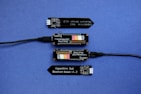 A series of electronic components displayed on a blue surface, including two black modules labeled 'Plant Wing' with colorful bar graphics and USB connectors. Two black capacitive soil moisture sensors version 1.2 are also present, with one shown connected via black cables.