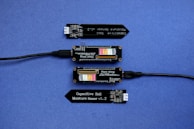 A series of electronic components displayed on a blue surface, including two black modules labeled 'Plant Wing' with colorful bar graphics and USB connectors. Two black capacitive soil moisture sensors version 1.2 are also present, with one shown connected via black cables.