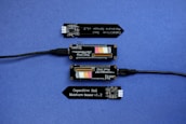 A series of electronic components displayed on a blue surface, including two black modules labeled 'Plant Wing' with colorful bar graphics and USB connectors. Two black capacitive soil moisture sensors version 1.2 are also present, with one shown connected via black cables.