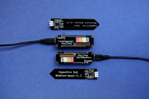 A series of electronic components displayed on a blue surface, including two black modules labeled 'Plant Wing' with colorful bar graphics and USB connectors. Two black capacitive soil moisture sensors version 1.2 are also present, with one shown connected via black cables.