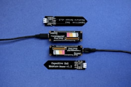 A series of electronic components displayed on a blue surface, including two black modules labeled 'Plant Wing' with colorful bar graphics and USB connectors. Two black capacitive soil moisture sensors version 1.2 are also present, with one shown connected via black cables.