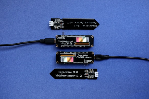 A series of electronic components displayed on a blue surface, including two black modules labeled 'Plant Wing' with colorful bar graphics and USB connectors. Two black capacitive soil moisture sensors version 1.2 are also present, with one shown connected via black cables.