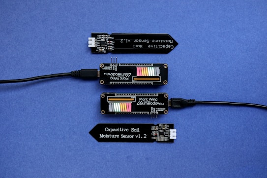 A series of electronic components displayed on a blue surface, including two black modules labeled 'Plant Wing' with colorful bar graphics and USB connectors. Two black capacitive soil moisture sensors version 1.2 are also present, with one shown connected via black cables.