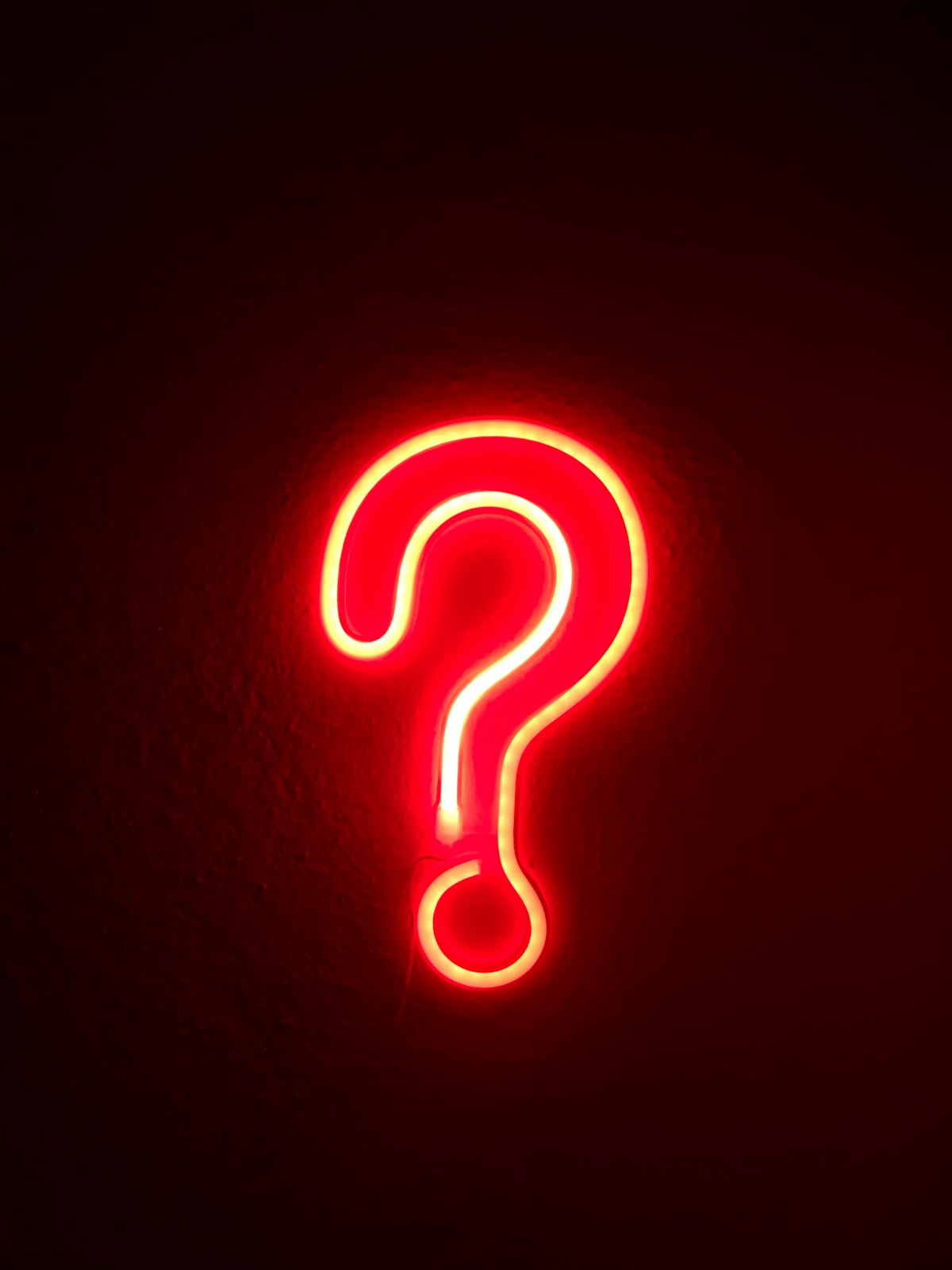 A red neon outlined question mark against a black wall.  The neon There is a red glow against the white and yellow outline of the question mark.