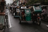 A line of tricycles travels down a narrow, wet street. Buildings and shops flank both sides, with people and electrical wires visible above. The tricycles are motorized, with some having passengers seated inside.