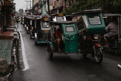 A line of tricycles travels down a narrow, wet street. Buildings and shops flank both sides, with people and electrical wires visible above. The tricycles are motorized, with some having passengers seated inside.