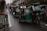 A line of tricycles travels down a narrow, wet street. Buildings and shops flank both sides, with people and electrical wires visible above. The tricycles are motorized, with some having passengers seated inside.