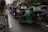 A line of tricycles travels down a narrow, wet street. Buildings and shops flank both sides, with people and electrical wires visible above. The tricycles are motorized, with some having passengers seated inside.