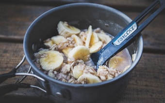 A metal pot containing oatmeal mixed with sliced bananas is placed on a wooden surface. A black utensil labeled 'FORCLAZ' is resting inside the pot, used for stirring or serving the meal.