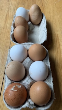 A dozen eggs in various shades of brown and white are placed in two cardboard egg cartons on a wooden surface. Some eggs have slight textures or marks on their shells.