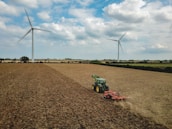 A large agricultural field with two wind turbines in the background. A green tractor is working on the field, which appears dry and harvested. The sky is partly cloudy, creating a serene atmosphere.