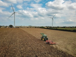 A large agricultural field with two wind turbines in the background. A green tractor is working on the field, which appears dry and harvested. The sky is partly cloudy, creating a serene atmosphere.