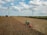 A large agricultural field with two wind turbines in the background. A green tractor is working on the field, which appears dry and harvested. The sky is partly cloudy, creating a serene atmosphere.