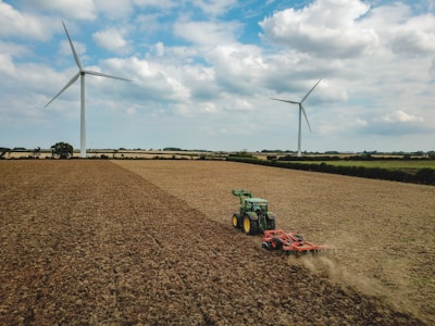 A large agricultural field with two wind turbines in the background. A green tractor is working on the field, which appears dry and harvested. The sky is partly cloudy, creating a serene atmosphere.