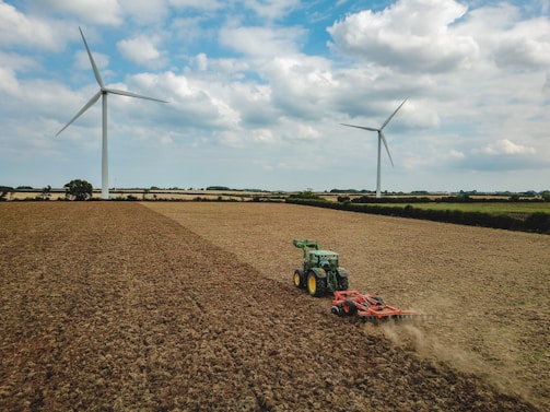 A large agricultural field with two wind turbines in the background. A green tractor is working on the field, which appears dry and harvested. The sky is partly cloudy, creating a serene atmosphere.