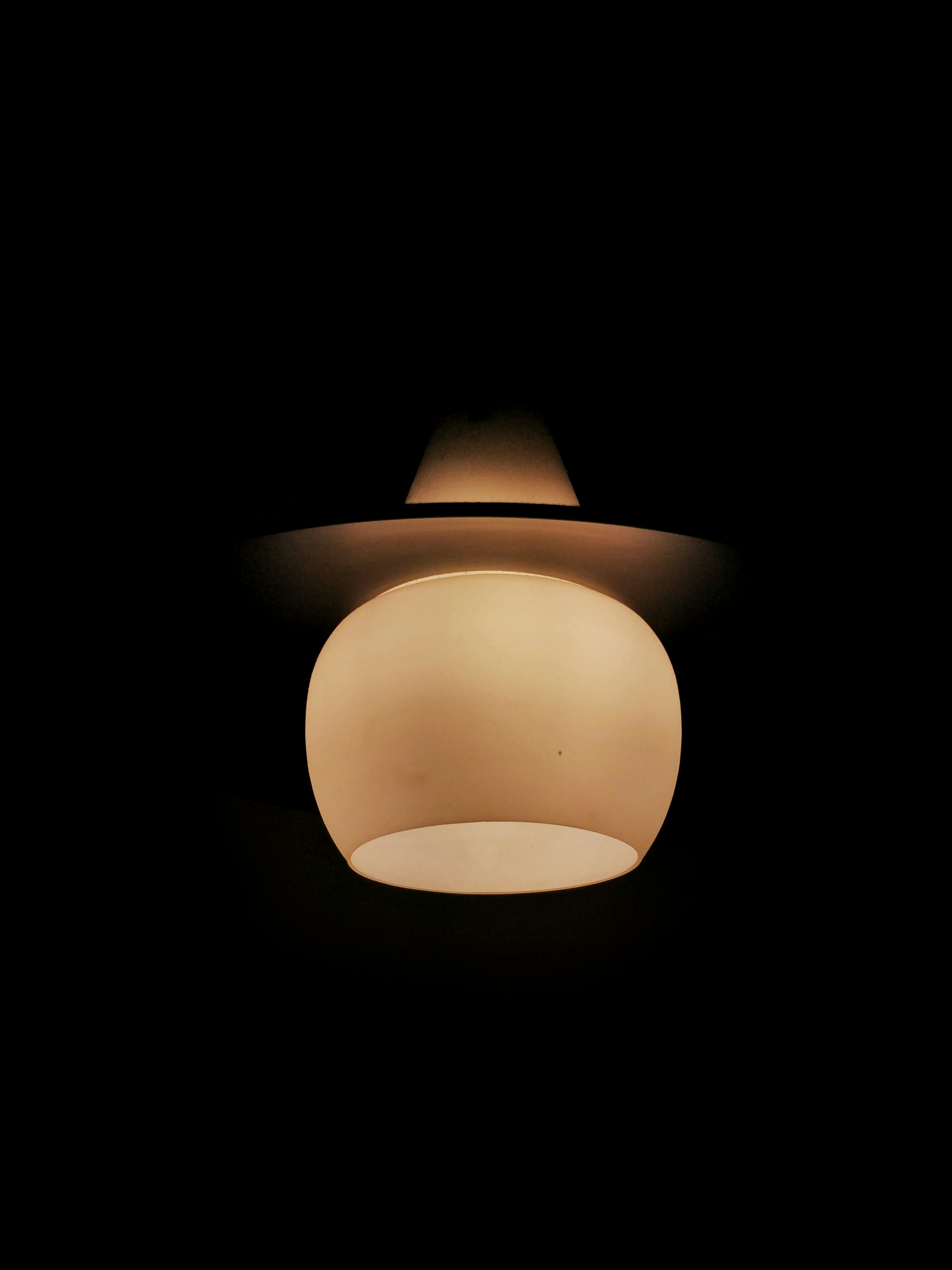 A ceiling light fixture with a simple, modern design is illuminated against a dark background. The light emanates a warm, soft glow, highlighting the contours of the fixture.