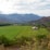A lush green agricultural field stretches across the landscape, bordered by dense forests and overshadowed by distant, rugged mountains. A solitary wooden barn sits in the middle of the field, adding a rustic touch to the serene setting under a partly cloudy sky.