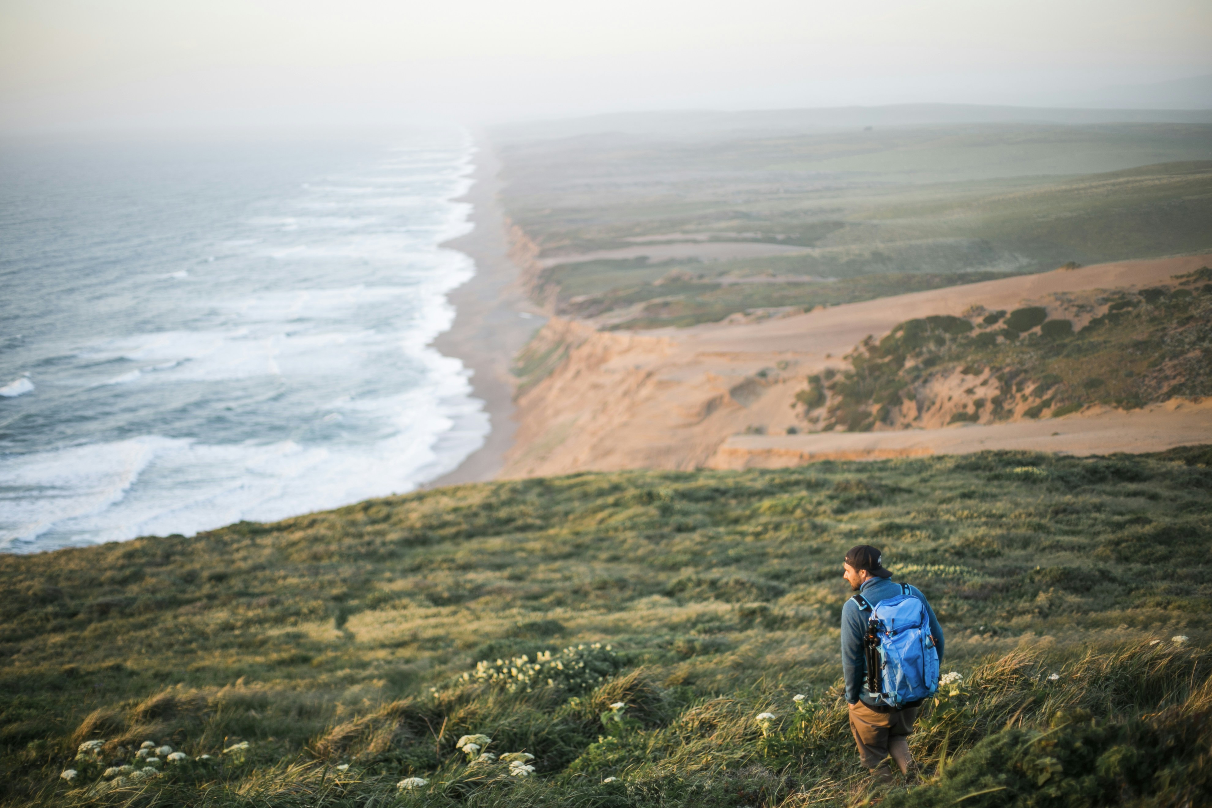 man in blue jacket standing on green grass field near body of water during daytime, A man wearing a blue backpack is standing in a grassy field looking towards the waves of the Pacific Ocean crashing on the beach in the Point Reyes National Seashore on the California coast.