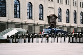 A row of riot police officers stands in front of a large, classical-style building with arched windows. The officers are equipped with shields and helmets and appear to be blocking access to the building. A large vehicle, which looks like a water cannon truck, is parked behind the officers. One person in front is holding a flag. The scene seems to be set in an urban area.