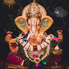 A vibrant and detailed idol of Lord Ganesha adorned with a golden crown and intricate jewelry. The deity holds a pink lotus flower in the left hand, symbolizing spiritual enlightenment. The idol is decorated with colorful garlands and intricate patterns, with a shiny metallic object in the right hand. The dark background with floral elements accentuates the bright and festive appearance of the idol.