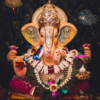 A vibrant and detailed idol of Lord Ganesha adorned with a golden crown and intricate jewelry. The deity holds a pink lotus flower in the left hand, symbolizing spiritual enlightenment. The idol is decorated with colorful garlands and intricate patterns, with a shiny metallic object in the right hand. The dark background with floral elements accentuates the bright and festive appearance of the idol.