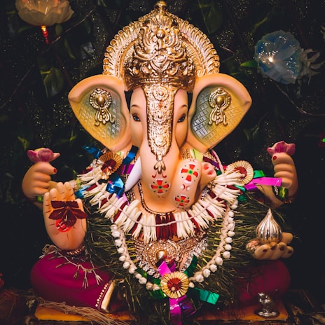 A vibrant and detailed idol of Lord Ganesha adorned with a golden crown and intricate jewelry. The deity holds a pink lotus flower in the left hand, symbolizing spiritual enlightenment. The idol is decorated with colorful garlands and intricate patterns, with a shiny metallic object in the right hand. The dark background with floral elements accentuates the bright and festive appearance of the idol.