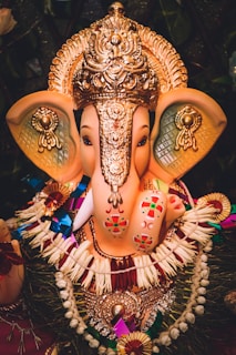 An elaborately decorated idol of the elephant-headed deity Ganesh, adorned with an ornate golden headdress and various colorful ornaments. The sculpture features intricate patterns and vibrant designs, with floral garlands and symbolic decorations enhancing its appearance.