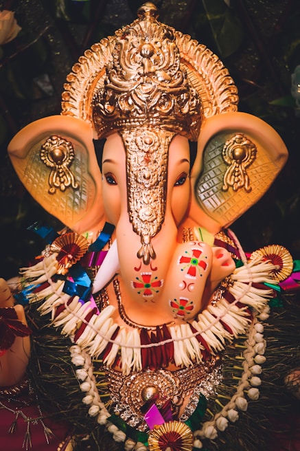 An elaborately decorated idol of the elephant-headed deity Ganesh, adorned with an ornate golden headdress and various colorful ornaments. The sculpture features intricate patterns and vibrant designs, with floral garlands and symbolic decorations enhancing its appearance.