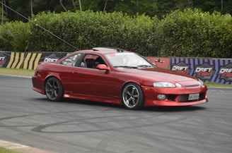 A red sports car is captured drifting on a race track with advertisements labeled 'Drift Fight' in the background. The car has tinted windows and an open driver's window. There are tire marks visible on the track, and the scene is surrounded by tall green hedges.