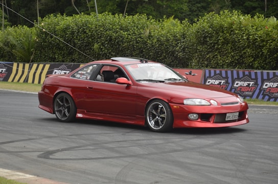 A red sports car is captured drifting on a race track with advertisements labeled 'Drift Fight' in the background. The car has tinted windows and an open driver's window. There are tire marks visible on the track, and the scene is surrounded by tall green hedges.
