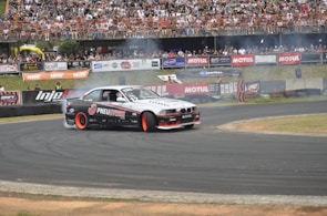 A drift car powers through a racetrack corner, with smoking tires and a crowd of spectators in the stands. The car has prominent branding and is surrounded by advertising banners.