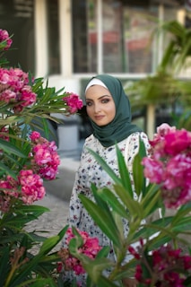 A woman wearing a green hijab and floral dress stands amidst vibrant pink flowers, with leafy greenery surrounding her. Behind her is a blurred background of a building with windows.