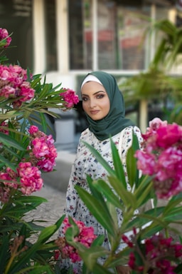A woman wearing a green hijab and floral dress stands amidst vibrant pink flowers, with leafy greenery surrounding her. Behind her is a blurred background of a building with windows.