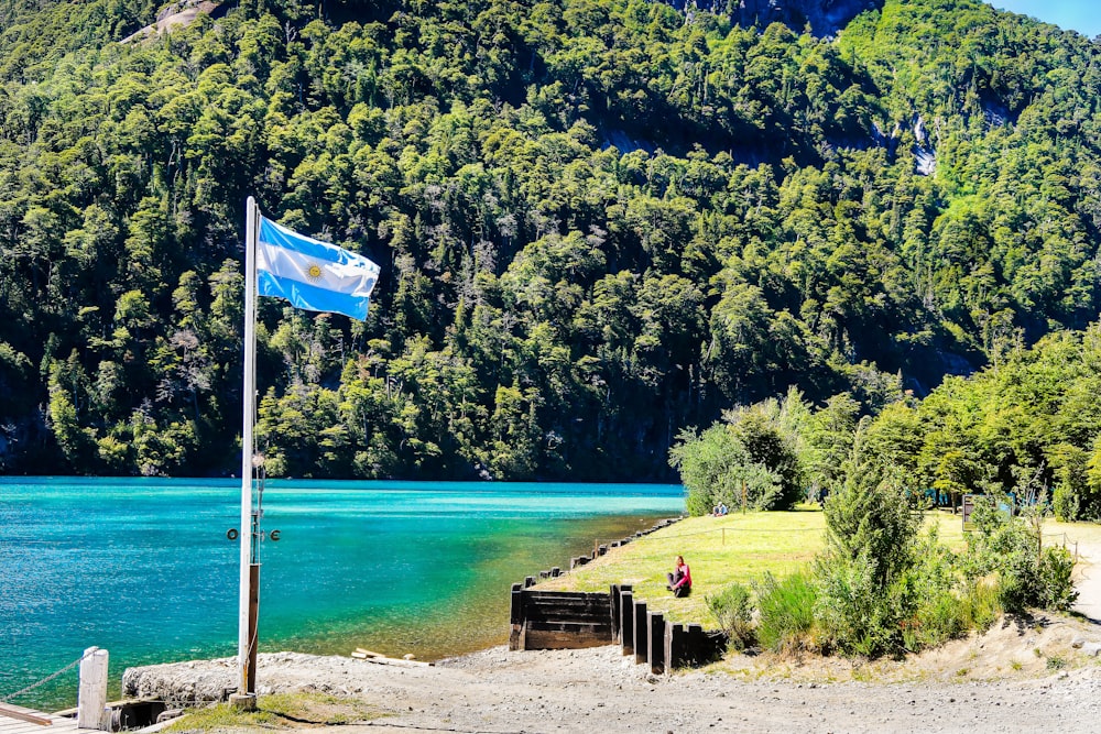 낮 동안 수역 근처의 갈색 나무 벤치에 파란색 깃발