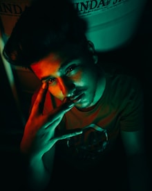 A person with styled hair illuminated by dramatic red and green lighting, making a hand gesture near their face. The setting is dark, with some text visible in the background.