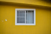 A white-framed window with two sliding glass panels is set in a bright yellow wall. The window is partially covered with sheer white curtains. The yellow wall has a rough texture and there are minor imperfections such as patches of peeling paint.