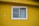 A white-framed window with two sliding glass panels is set in a bright yellow wall. The window is partially covered with sheer white curtains. The yellow wall has a rough texture and there are minor imperfections such as patches of peeling paint.
