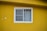 A white-framed window with two sliding glass panels is set in a bright yellow wall. The window is partially covered with sheer white curtains. The yellow wall has a rough texture and there are minor imperfections such as patches of peeling paint.