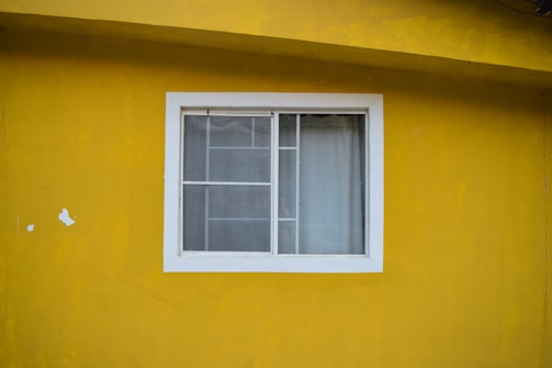 A white-framed window with two sliding glass panels is set in a bright yellow wall. The window is partially covered with sheer white curtains. The yellow wall has a rough texture and there are minor imperfections such as patches of peeling paint.