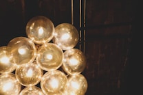 A cluster of glowing light bulbs arranged in a circular formation, set against a dark background with vertical beams. The bulbs emit a warm, golden light creating a cozy and ambient atmosphere.