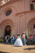 A young girl wearing a white dress stands in front of a large brick church with intricate design features. Surrounding her are groups of people, including families and children, some of whom are engaged in conversation or walking by. The atmosphere suggests a festive or ceremonial occasion, possibly a religious event.