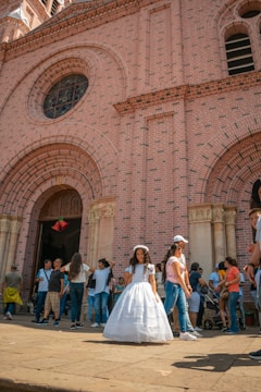 A young girl wearing a white dress stands in front of a large brick church with intricate design features. Surrounding her are groups of people, including families and children, some of whom are engaged in conversation or walking by. The atmosphere suggests a festive or ceremonial occasion, possibly a religious event.