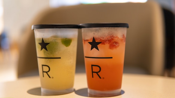 Two branded cups with black lids on a table, one containing a yellowish drink with lime and the other a reddish drink with fruit pieces.