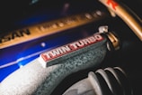 Close-up view of a metallic car engine component with the words 'TWIN TURBO' prominently displayed in red on a textured grey surface. Nearby, a blue surface and gold metallic elements feature subtly engraved markings, adding to the industrial aesthetic.