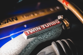 Close-up view of a metallic car engine component with the words 'TWIN TURBO' prominently displayed in red on a textured grey surface. Nearby, a blue surface and gold metallic elements feature subtly engraved markings, adding to the industrial aesthetic.
