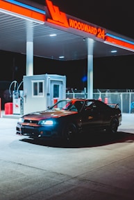A sleek black sports car is parked under the illuminated canopy of a gas station at night. The gas station sign reads 'WOODWARD 24' in bright red neon lights. The area is lit by overhead lights, creating a dramatic contrast of shadows and highlights. There's a small service booth nearby and various gas pumps visible in the background.