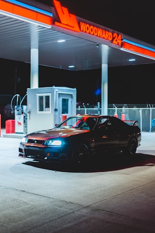 A sleek black sports car is parked under the illuminated canopy of a gas station at night. The gas station sign reads 'WOODWARD 24' in bright red neon lights. The area is lit by overhead lights, creating a dramatic contrast of shadows and highlights. There's a small service booth nearby and various gas pumps visible in the background.