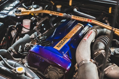 The image features a detailed view of a car engine, highlighting a blue Nissan engine cover with an inscription, various hoses, pipes, and a metallic frame with 'Skyline R' written on it. The engine components are clean and well-arranged, indicating a high-performance setup.