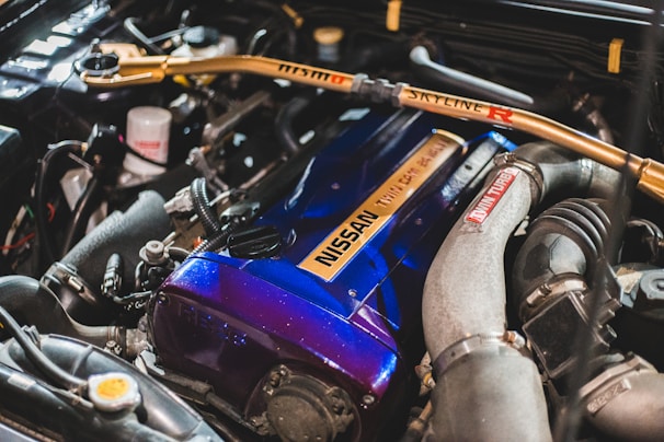 The image features a detailed view of a car engine, highlighting a blue Nissan engine cover with an inscription, various hoses, pipes, and a metallic frame with 'Skyline R' written on it. The engine components are clean and well-arranged, indicating a high-performance setup.